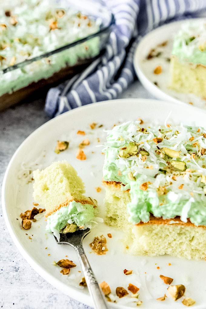 A forkful of cake next to a square piece of Watergate cake on a white plate with chopped pistachios and shredded coconut sprinkled around.  Cake slice on a plate and the whole baking dish with cake is in the background.