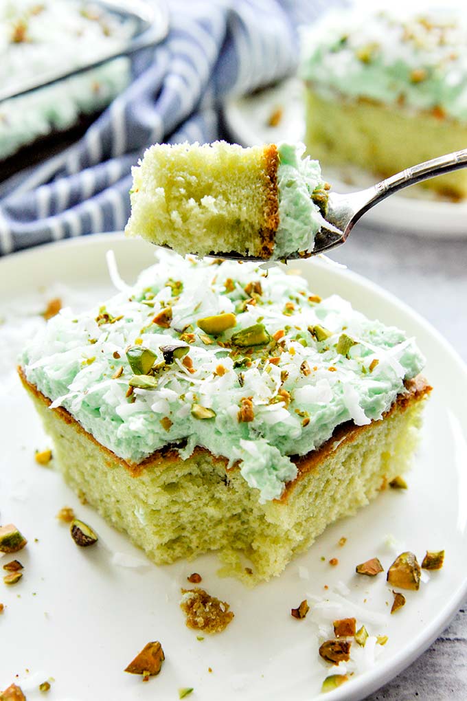 A forkful of cake above a square piece of Watergate cake on a white plate with chopped pistachios and shredded coconut sprinkled around.  Cake slice on a plate and the whole baking dish with cake is in the background.