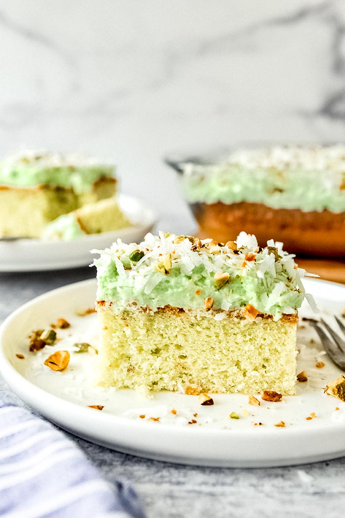 A square piece of Watergate cake on a white plate with chopped pistachios and shredded coconut sprinkled around. Cake slice on a plate and the whole baking dish with cake is in the background.