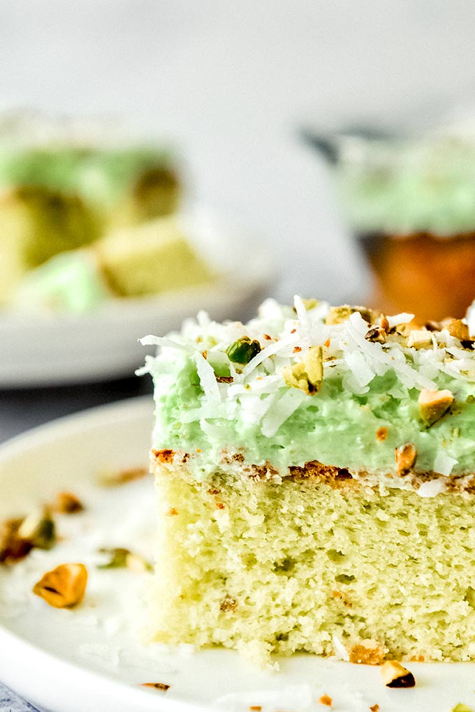 An up close view of a square piece of Watergate cake on a white plate with chopped pistachios and shredded coconut sprinkled around.  Cake slice on a plate and the whole baking dish with cake is in the background.