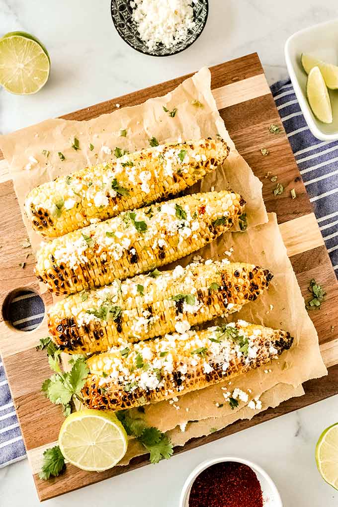 Four grilled ears of corn lined up spread with mayo, Cotija cheese, chili powder, and cilantro leaves on a cutting board.  Bowls of cut limes and cheese.