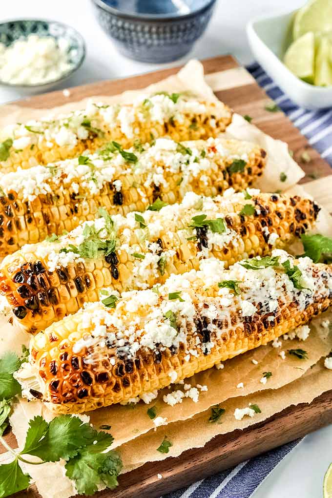 Four grilled ears of corn spread with mayo, Cotija cheese, chili powder, and cilantro leaves. Bowls of cut limes and cheese.
