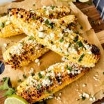 Four grilled ears of corn lined up spread with mayo, Cotija cheese, chili powder, and cilantro leaves. With a slice of lime.