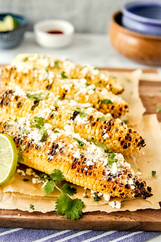Four grilled ears of corn spread with mayo, Cotija cheese, chili powder, and cilantro leaves.  Bowls of cut limes and cheese.