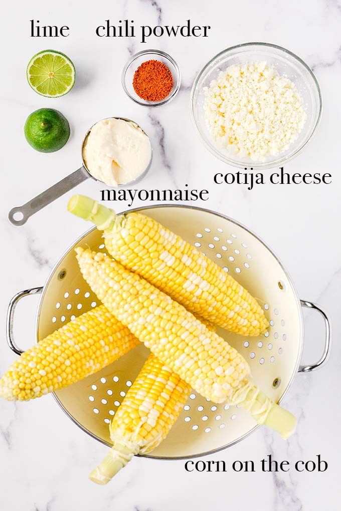 All of the ingredients to make Elotes Mexicanos like ears of corn, Cotija cheese, chili powder, mayonnaise, and lime.