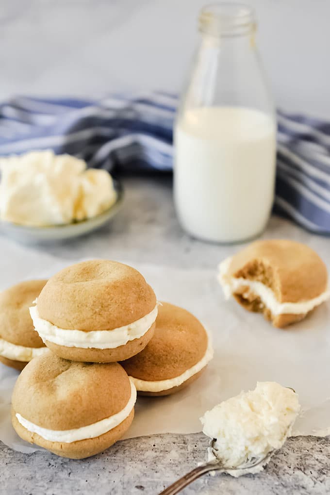 A pile of fluffernutter cookies stacked near a spoon of frosting.  A bowl of frosting, a bottle of milk, and a cookie with a bite missing in the background