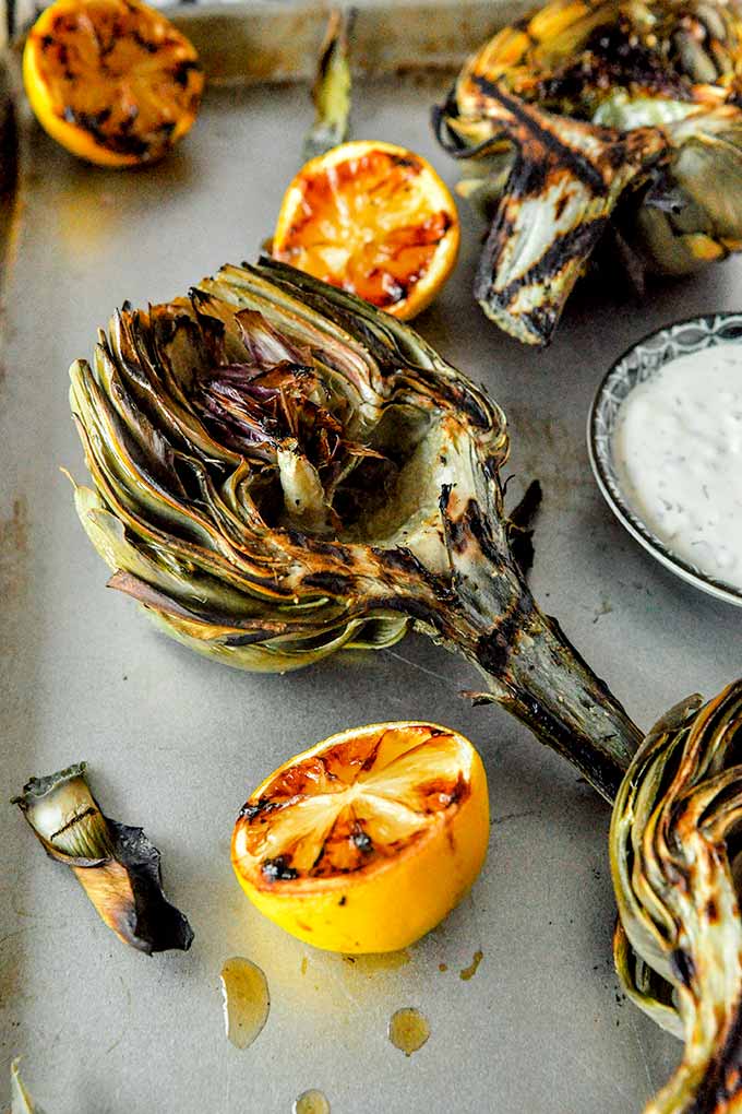 A grilled artichoke half with some grilled lemon halves and a small bowl of lemon aioli.