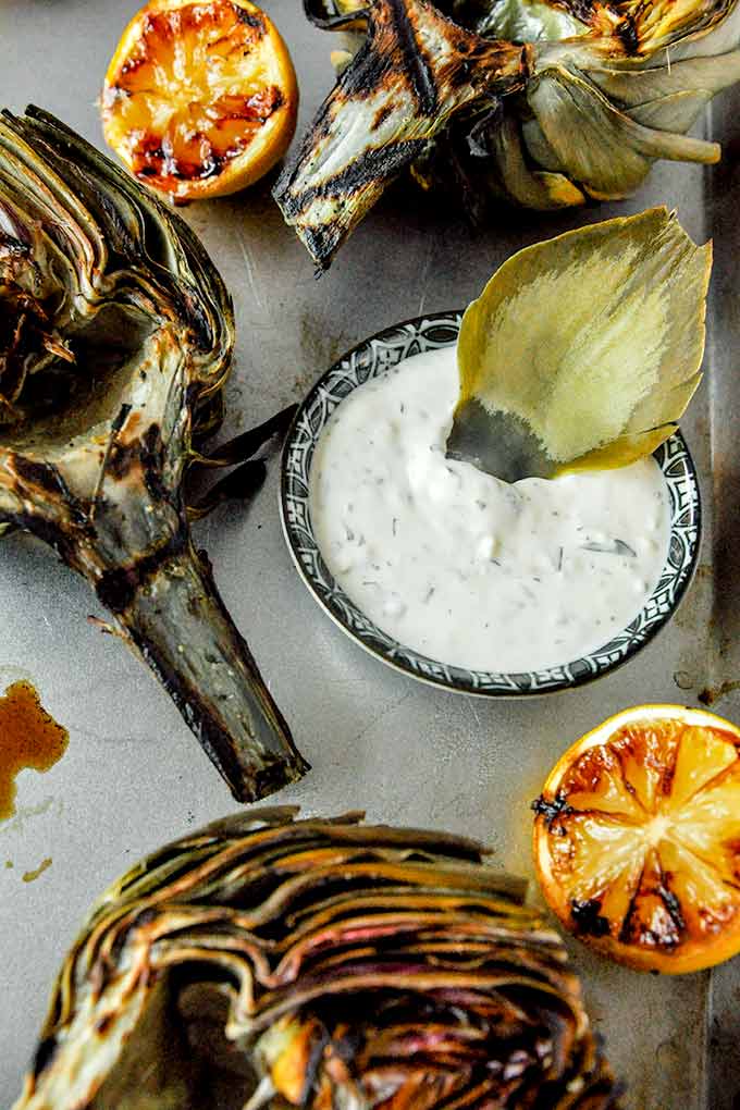 An artichoke leaf in the dipping sauce with a few grilled artichokes and lemon halves.