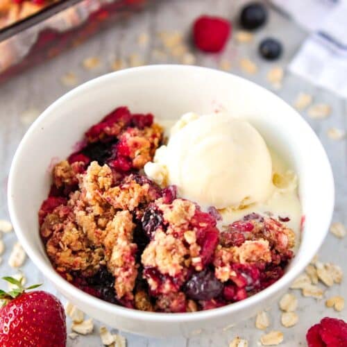 Triple berry crisp in a white bowl with a scoop of ice cream and baking pan of crisp in the background.
