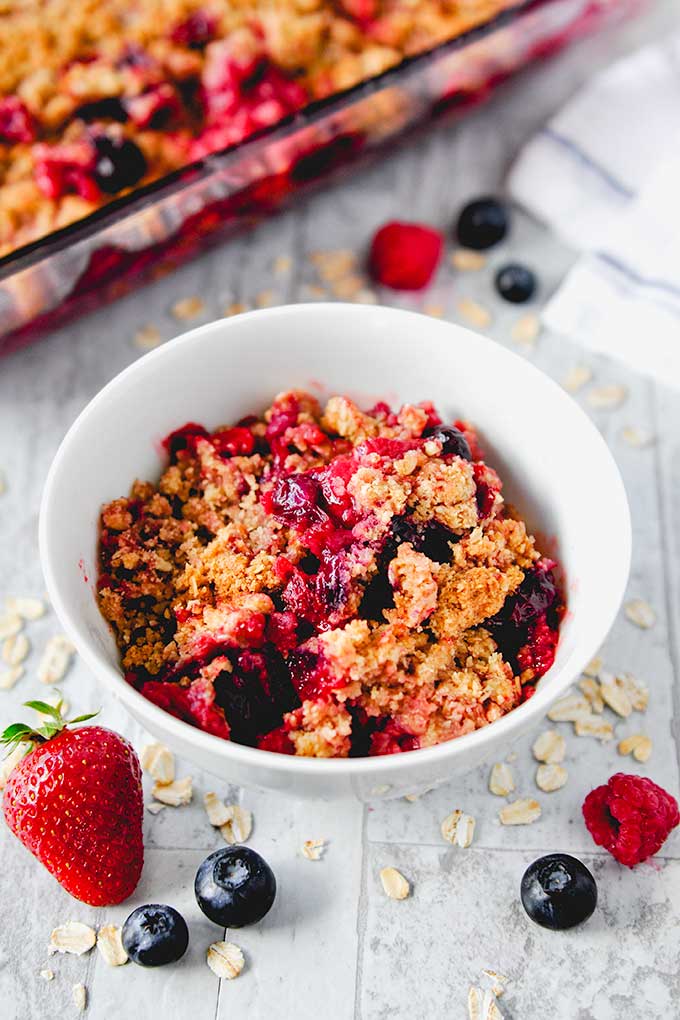 Triple berry crisp in a white bowl with berries and baking pan of crisp in the background.