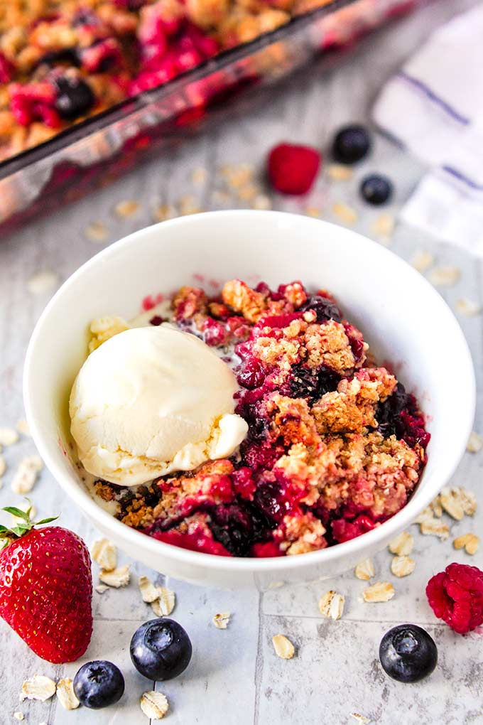 Triple berry crisp in a white bowl with a scoop of ice cream and baking pan of crisp in the background.