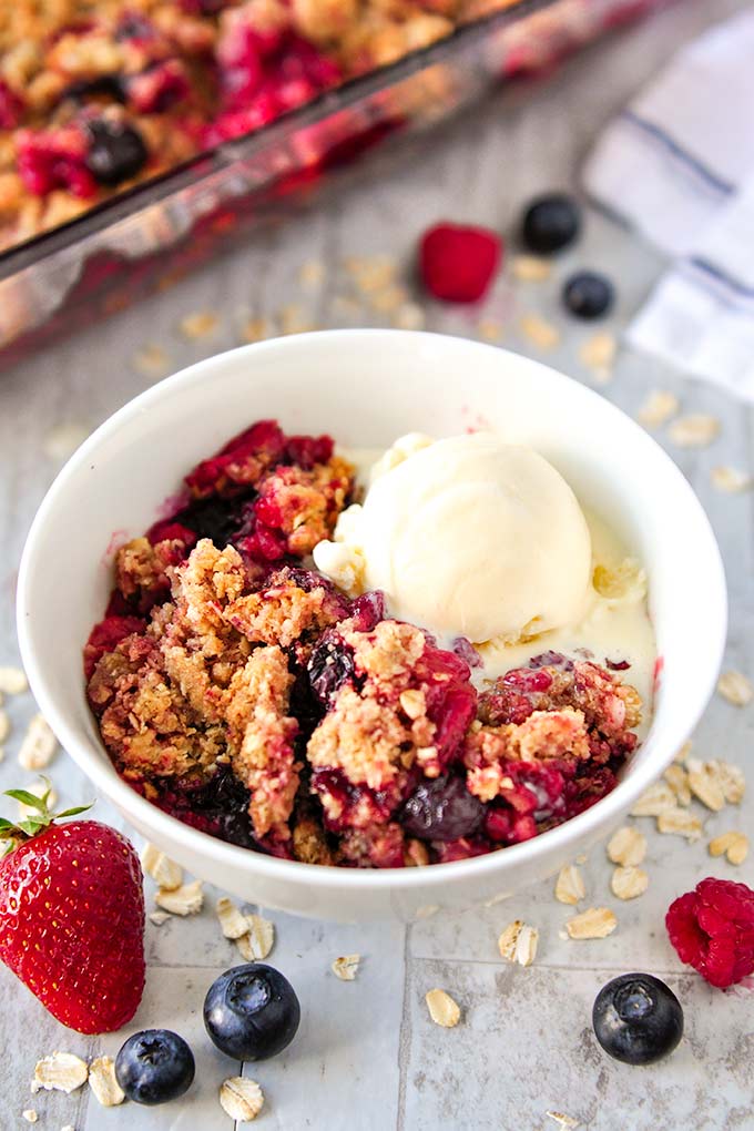 Triple berry crisp in a white bowl with a scoop of ice cream and baking pan of crisp in the background.