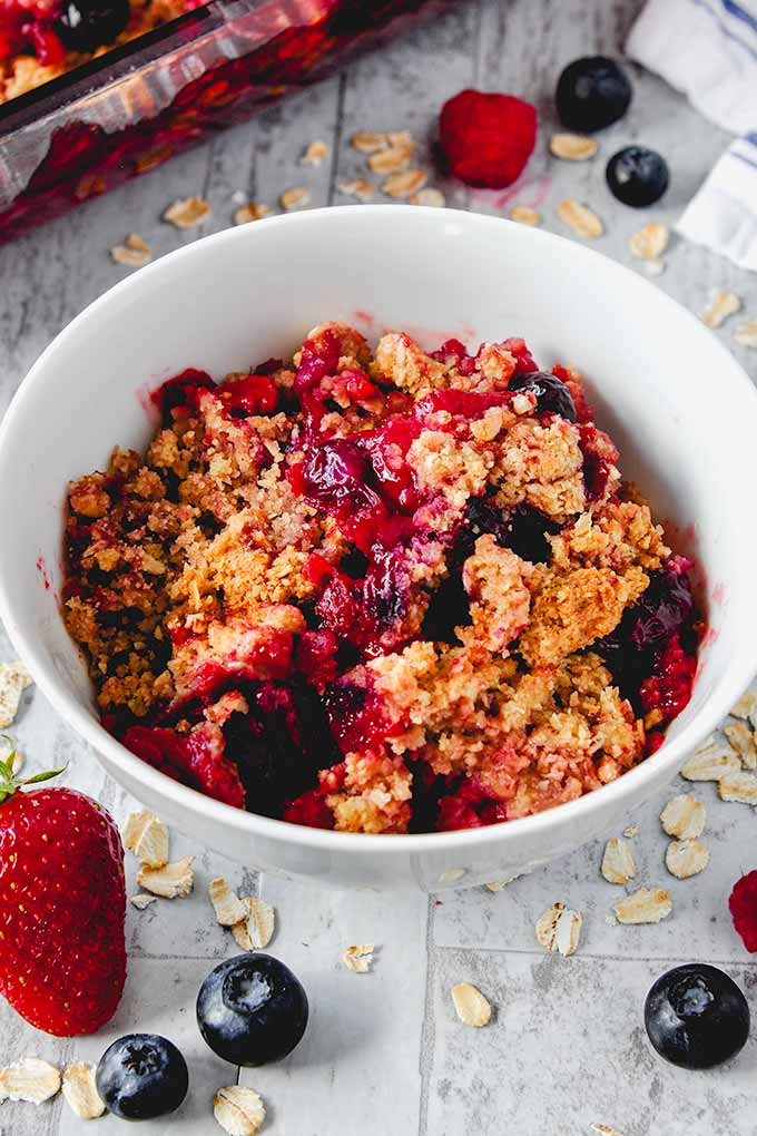 Triple berry crisp in a white bowl with berries in the front and baking pan of crisp in the background.