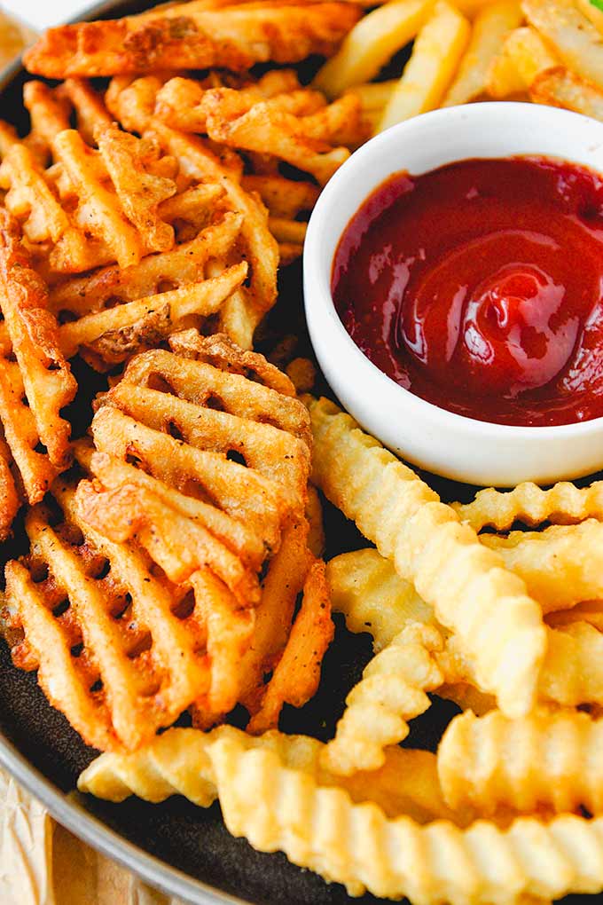A close up of waffle fries, crinkle fries, and straight fries with a cup of ketchup.