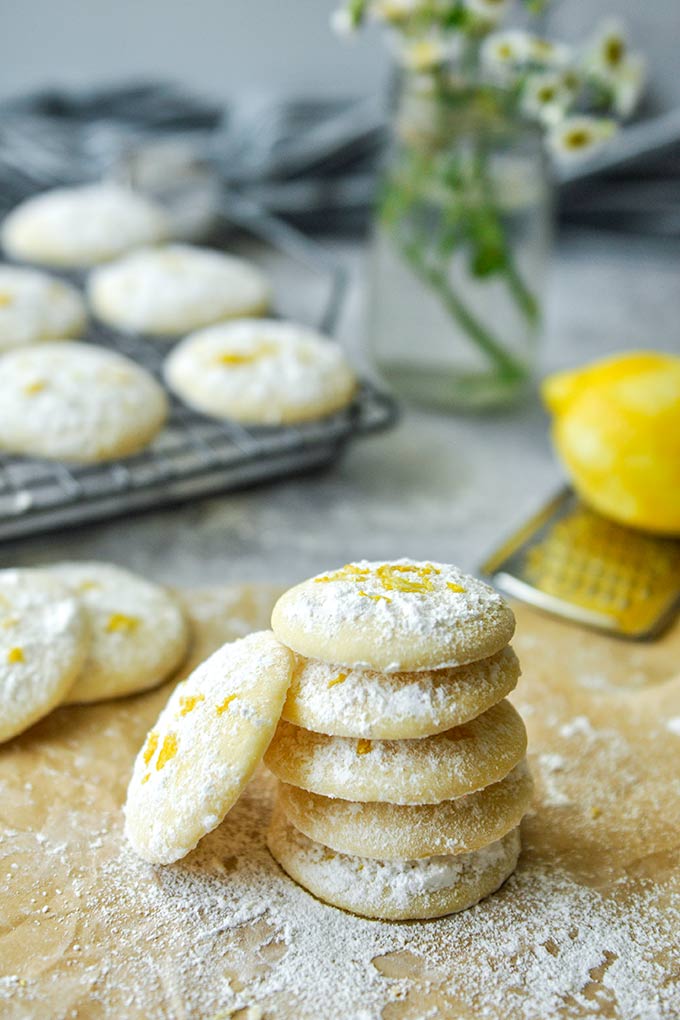 Lemon cooler cookies scattered around on parchment paper, a zested lemon on a grater, and a baking sheet of cookies