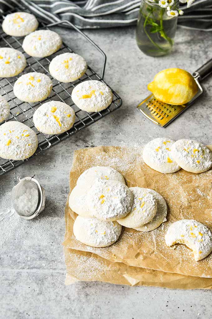 Lemon cooler cookies scattered around on parchment paper, a zested lemon on a grater, and a baking sheet of cookies