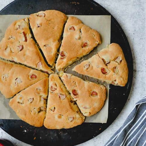 A whole ring of rhubarb scones placed together on a pizza stone with a red coffee cup and black and white striped towel.
