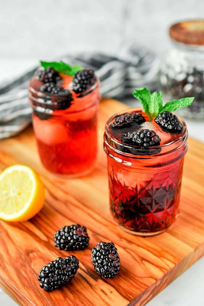 Two cups of blackberry iced tea with mint sprigs on a wood cutting board with a cut lemon and some blackberries.