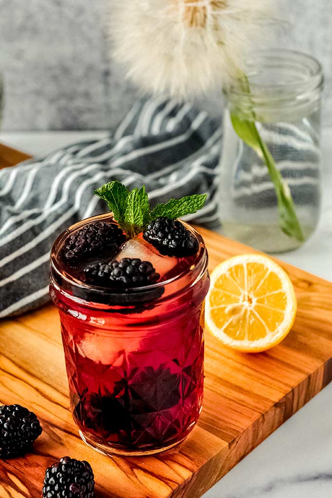 A cup of blackberry iced tea with mint sprigs on a wood cutting board with a cut lemon and some blackberries.
