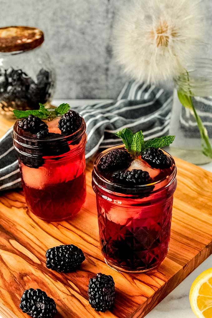 Two cups of blackberry iced tea with mint sprigs on a wood cutting board with a cut lemon and some blackberries.