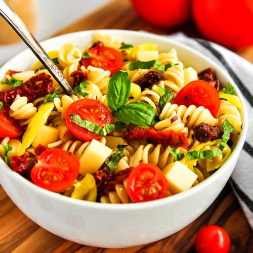 A bowl of spicy pasta salad topped with basil leaves, sun-dried tomatoes, fresh tomatoes, pepper flakes, and pepperoncinis.
