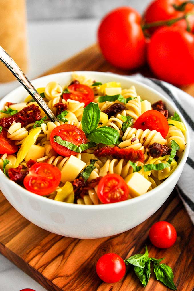 A bowl of spicy pasta salad topped with basil leaves, sun-dried tomatoes, fresh tomatoes, pepper flakes, and pepperoncinis.