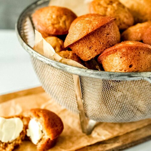 Mini banana muffins piled in a wire basket on a wooden cutting board. One buttered cut muffin next to basket.