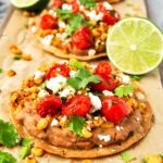 Three bean tostadas topped with baked corn and tomatoes, queso fresco and fresh lime halves.