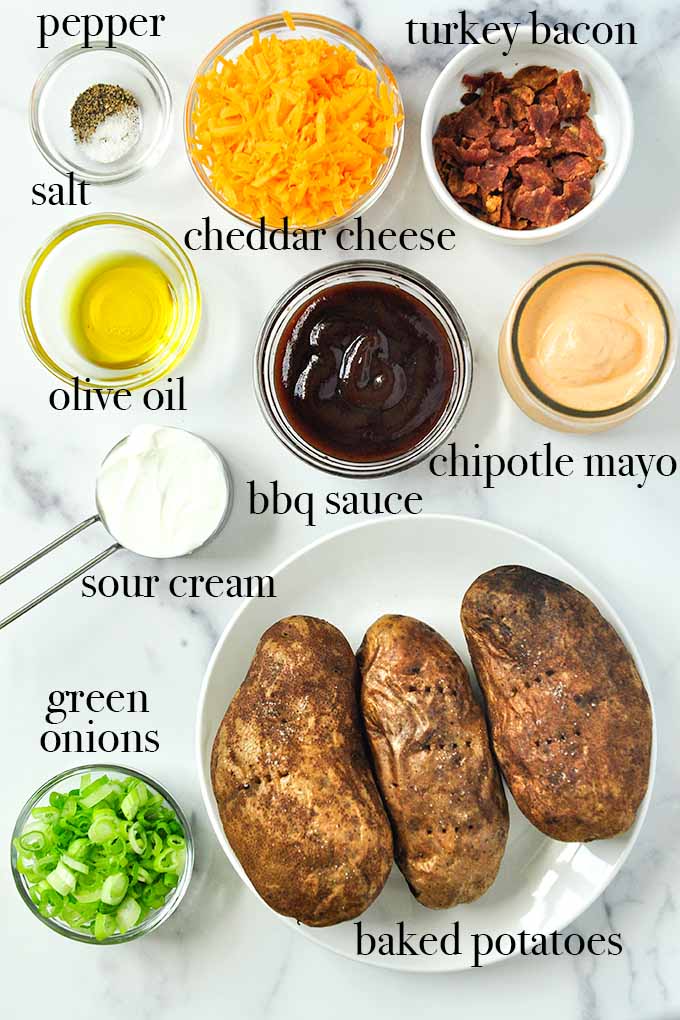 All of the ingredients needed to make potato skins including cheddar cheese, turkey bacon, bbq sauce, baked potatoes, and chipotle mayonnaise.