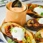 Air fryer potato skins topped with sour cream and green onions on a plate and a jar of chipotle mayonnaise with a spoon sticking out of it.