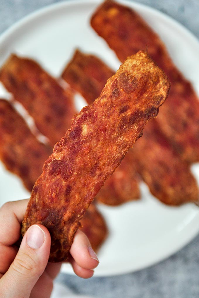 A piece of crispy turkey bacon being held above a plate of bacon.