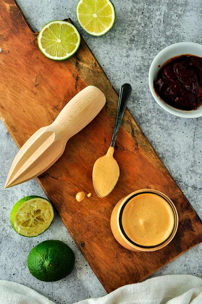 A cutting board with a jar of chipotle mayonnaise, a spoon by it's side covered in sauce.  Some limes lay about with a wooden reamer and a bowl of chipotle peppers.