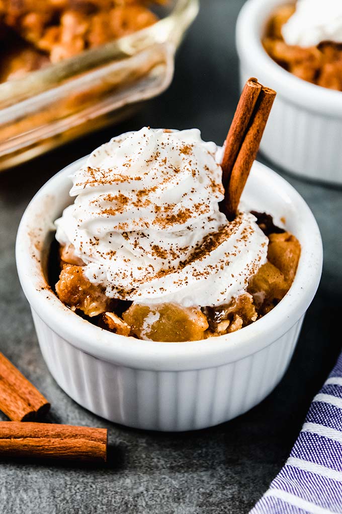 A small bowl of baked apple crisp topped with whipped cream, a sprinkling of cinnamon, and a cinnamon stick.
