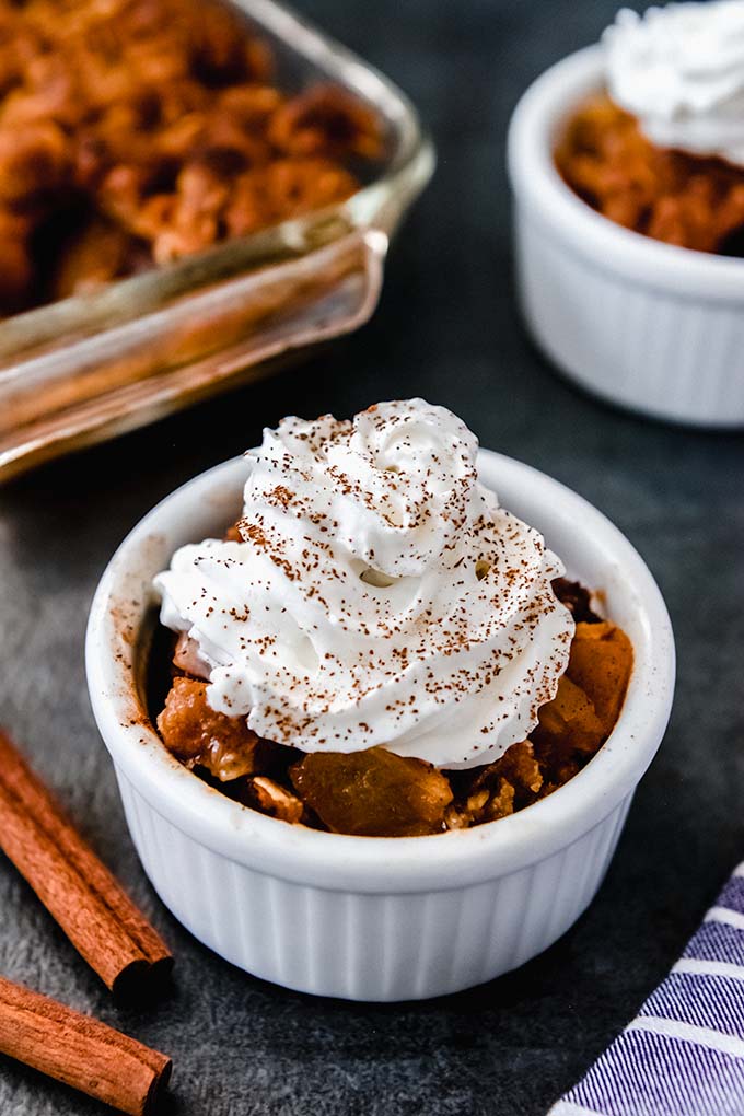 A small bowl of apple crisp with gala apples topped with whipped cream and a sprinkling of cinnamon beside cinnamon sticks. The baking dish of crisp is in the background.