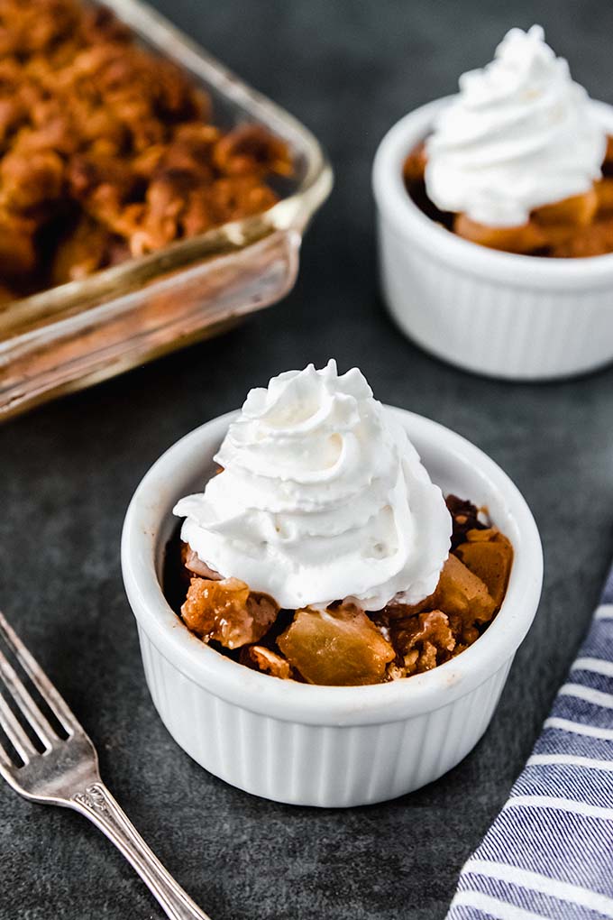 A small bowl of apple crisp with gala apples topped with whipped cream and a fork. A baking dish full of baked apples and another bowl of apple crisp in background.
