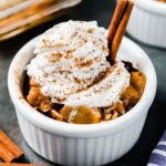 A small bowl of baked apple crisp topped with whipped cream, a sprinkling of cinnamon, and a cinnamon stick.