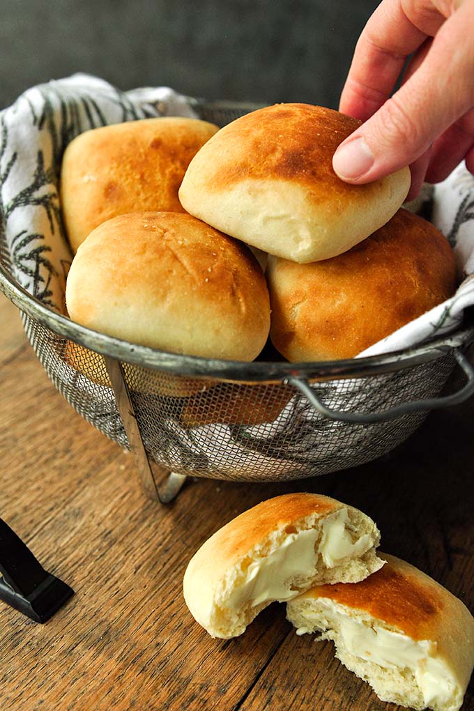 A hand reaching in to grab a roll from a basket with a buttered roll in front.