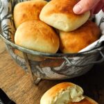 A hand reaching in to grab a roll from a basket with a buttered roll in front.