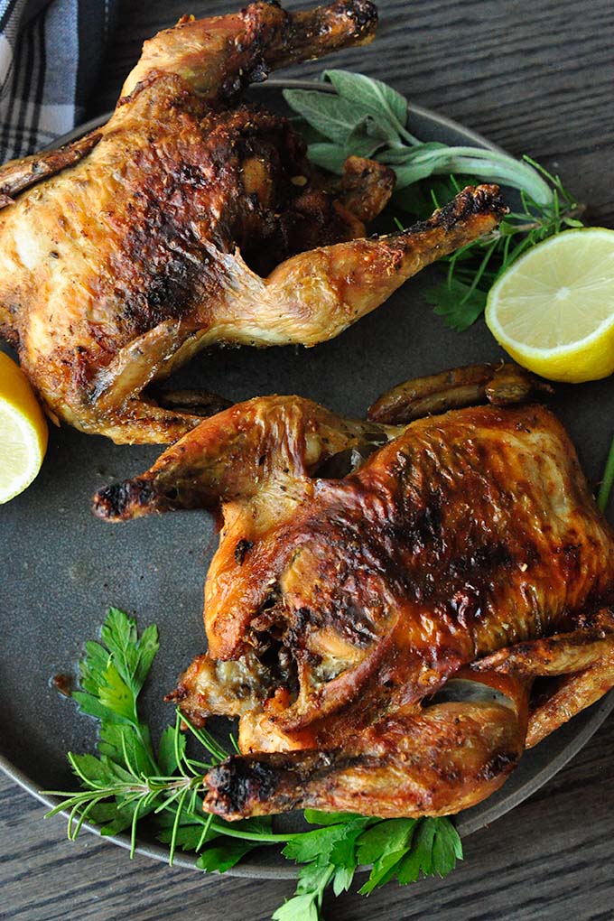 Two air fryer cornish hens on a plate with lemon halves and herbs.