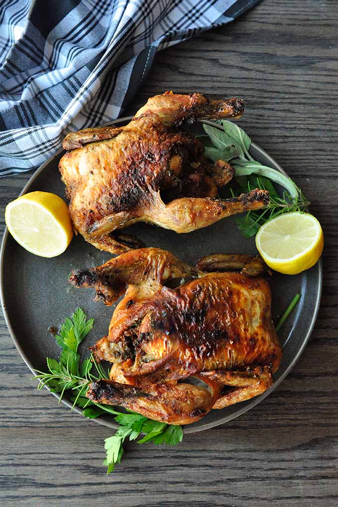 Two air fryer cornish hens on a plate with lemon halves and greens.