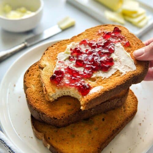A hand reaching into a stack of buttered and jelly toast on a plate.