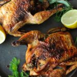 Two air fryer cornish hens on a plate with lemon halves and greens.