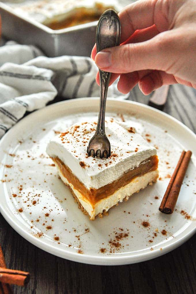 A square of no bake pumpkin pie cheesecake on a plate with cinnamon sticks and a fork sticking about to dig in.