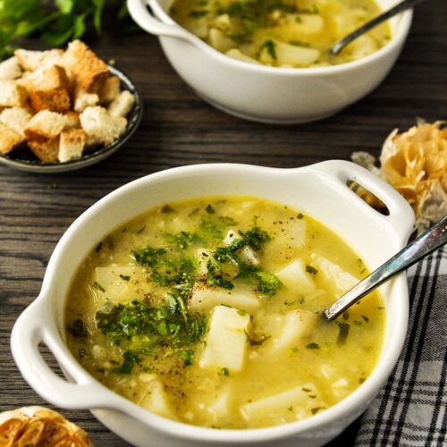 Two bowls of garlic potato soup with croutons, fresh parsley, and roasted garlic around.