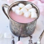 A metal cup full of strawberry hot chocolate, topped with marshmallows and pink towel in the background.