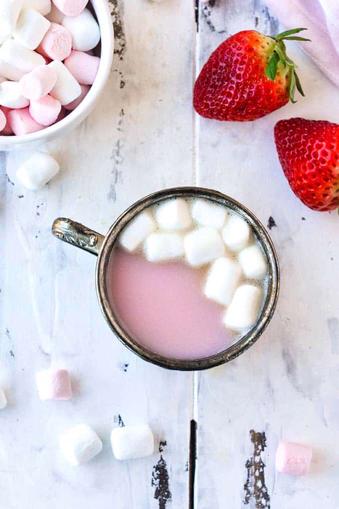 An above view of a metal cup full of strawberry hot cocoa topped with marshmallows.  A bowl of marshmallows and two strawberries are in the background.