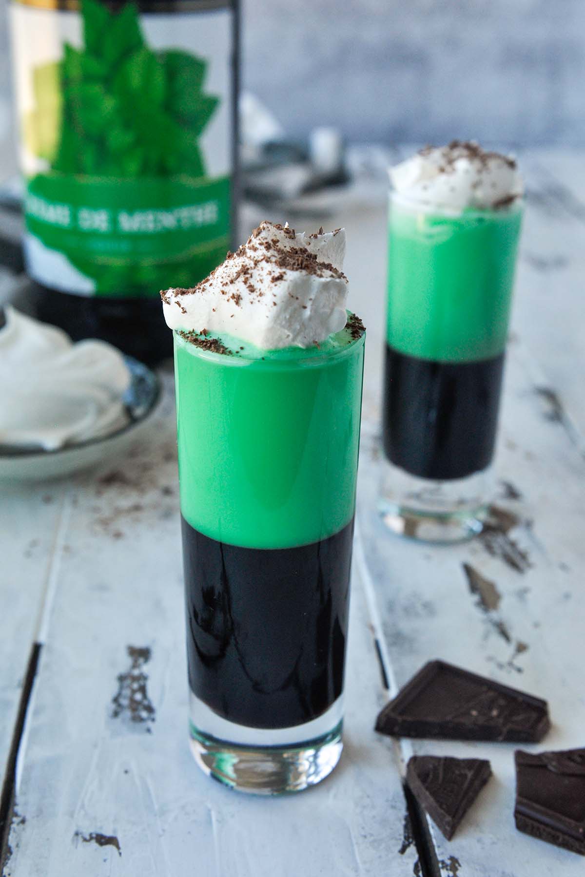 Two grasshopper shots topped with whipped cream and chocolate shavings with a bottle of Creme de menthe.