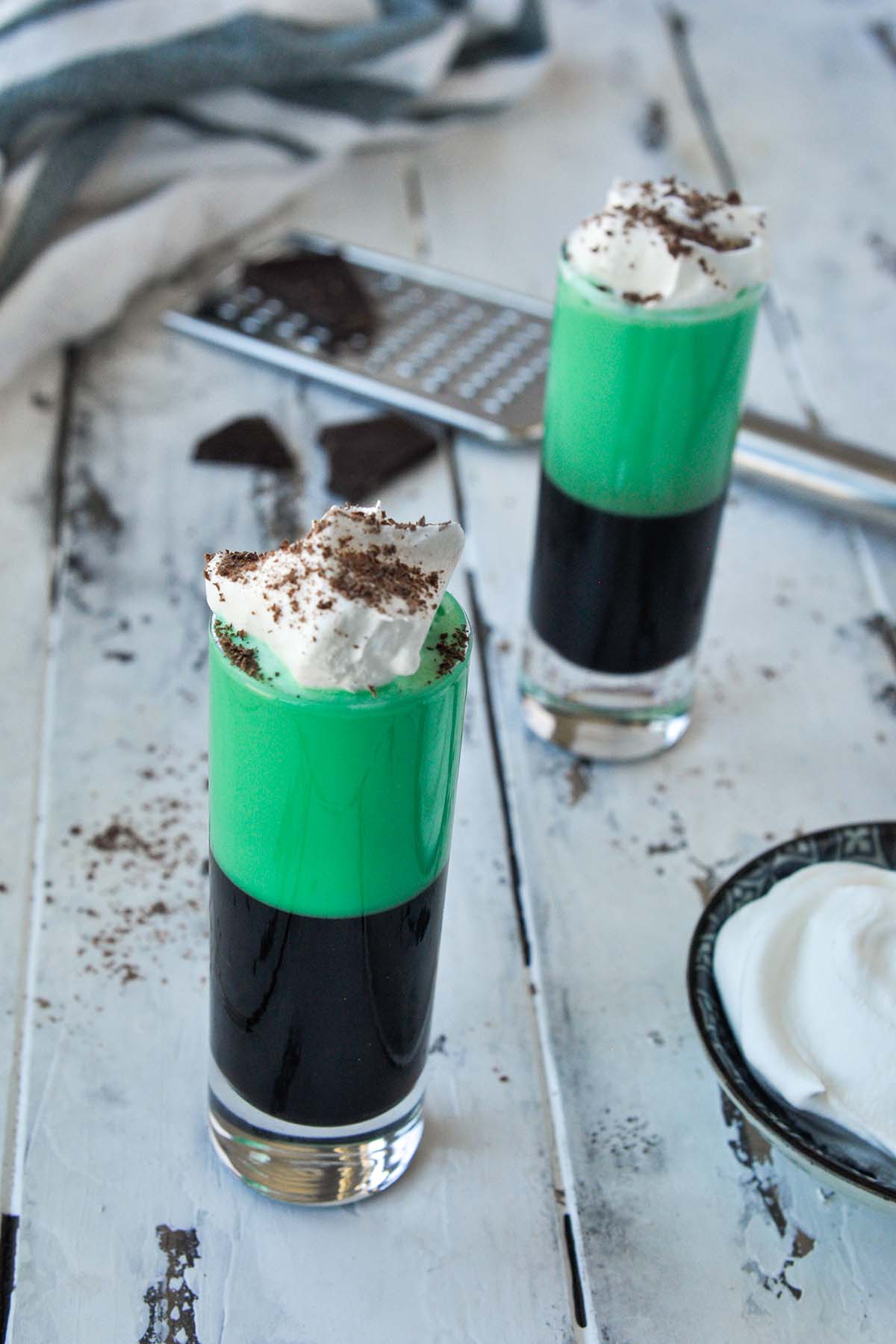 Two grasshopper shots topped with whipped cream and chocolate shavings with a small bowl of whipped cream.