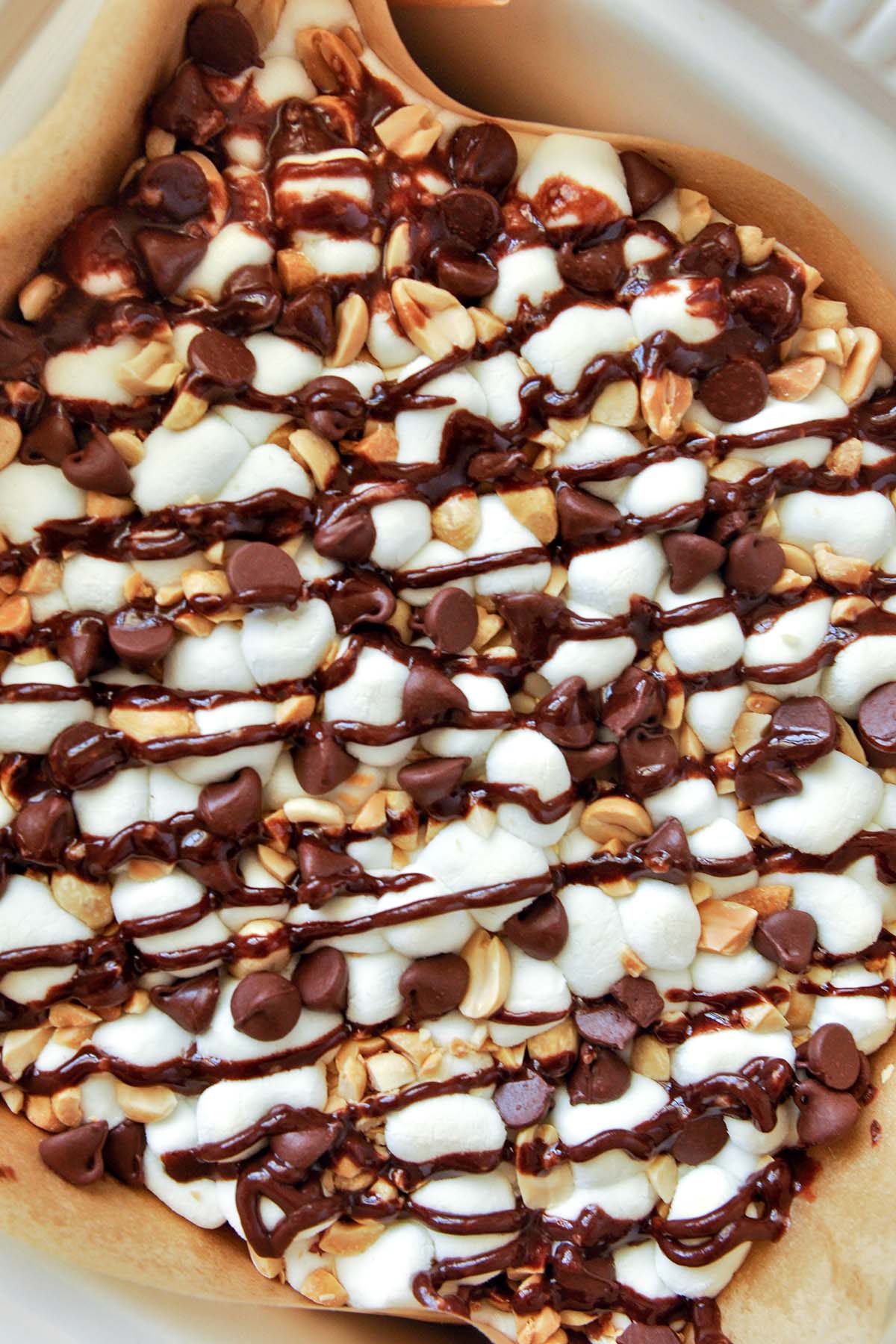 An above view of rocky road brownies topped with chocolate chips, marshmallows, peanuts, and a chocolate drizzle in a pan.