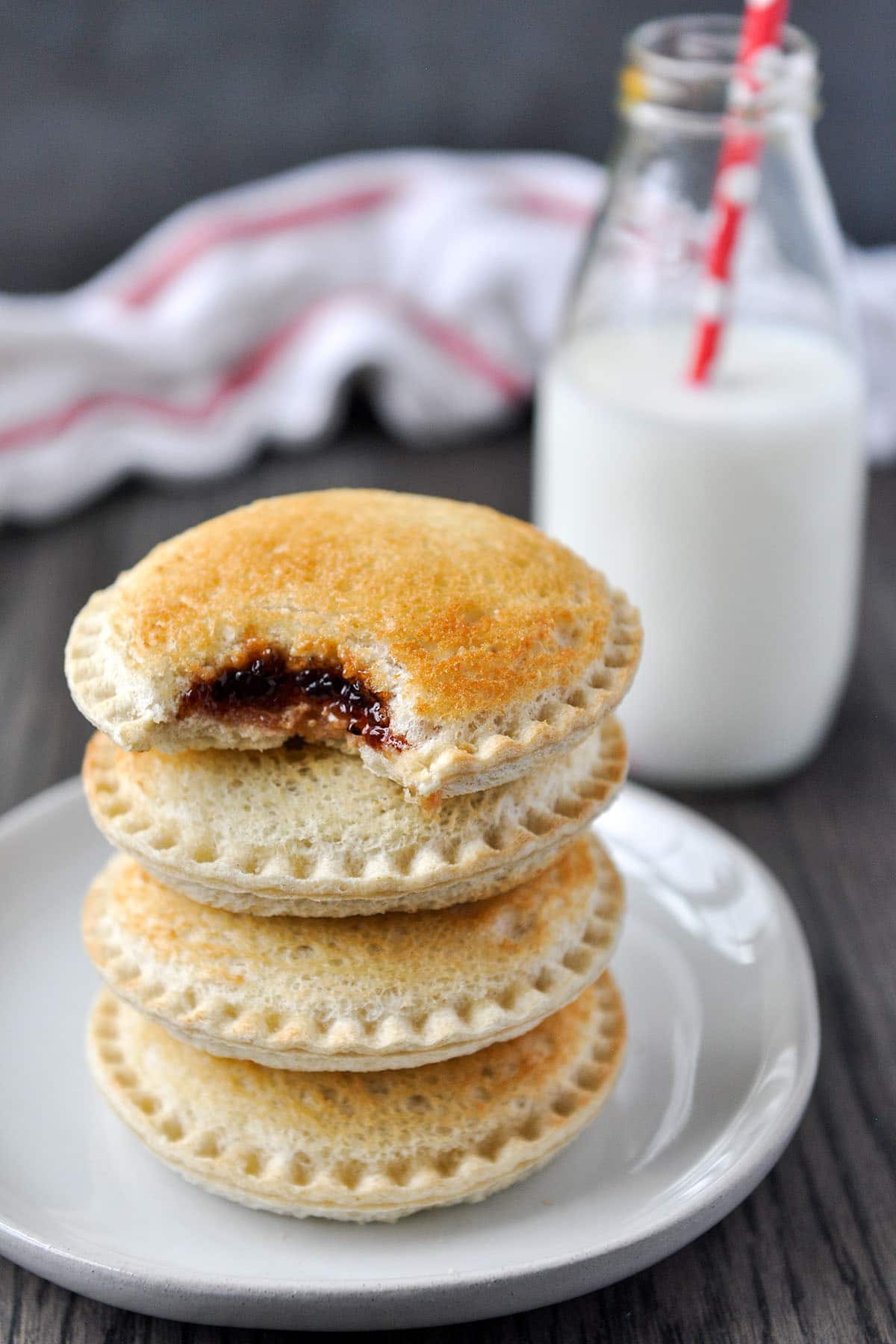 A stack of toasty Uncrustables on a plate, the top one is missing a bite and there is a glass bottle of milk and a white and red towel in the background.